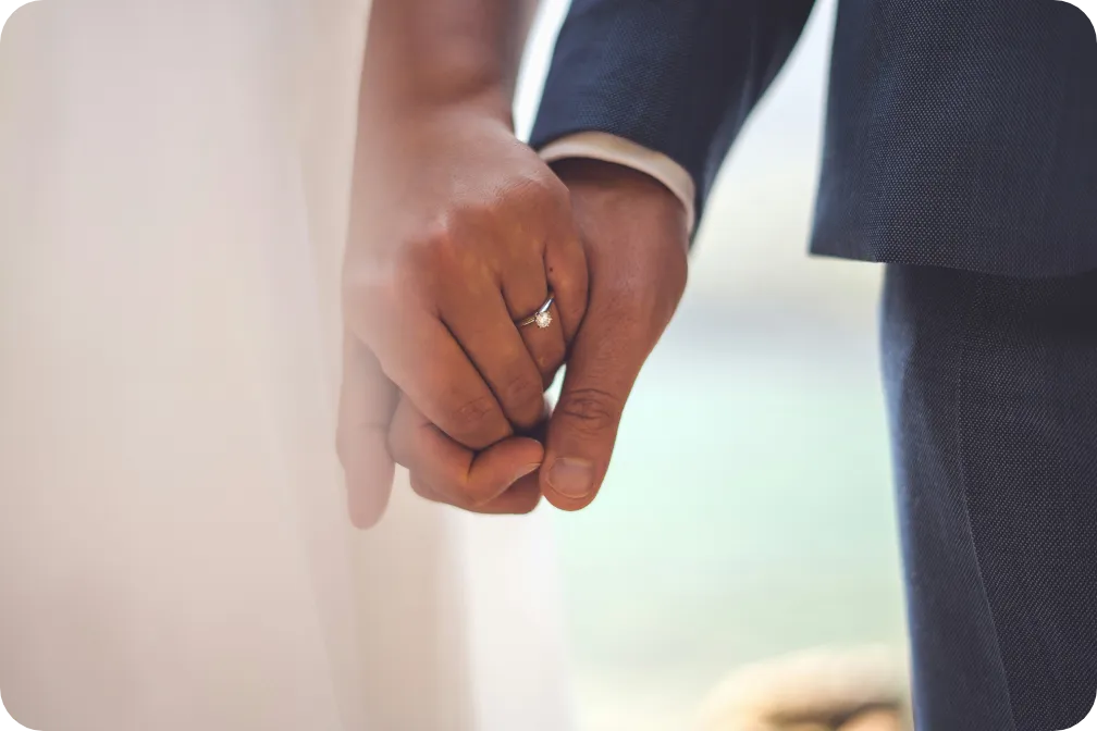 Ein Hochzeitspaar hält sich an den Händen. An der Hand der Braut ist ein Ring sichtbar.